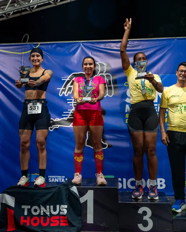 Any Britto, fundadora da Tonus House, no pódio de uma corrida de rua segurando o troféu, com outras duas corredoras e o banner da Tonus House no degrau